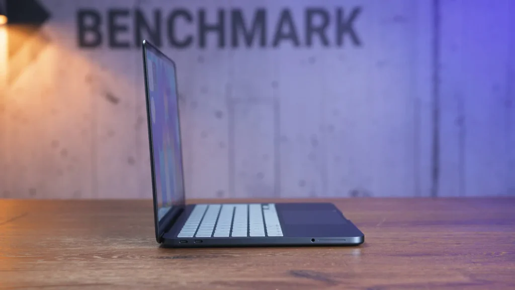 Laptop open on a wooden desk with a blurred wall reading BENCHMARK in the background.