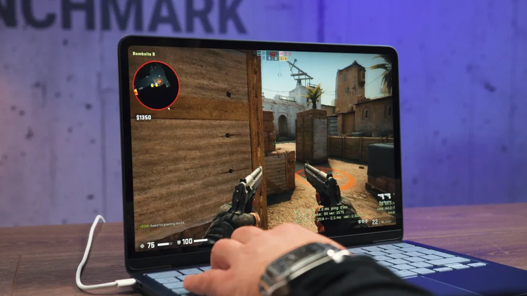 Hands on a laptop keyboard playing a first-person shooter game set in a desert map with dual pistols visible on screen.