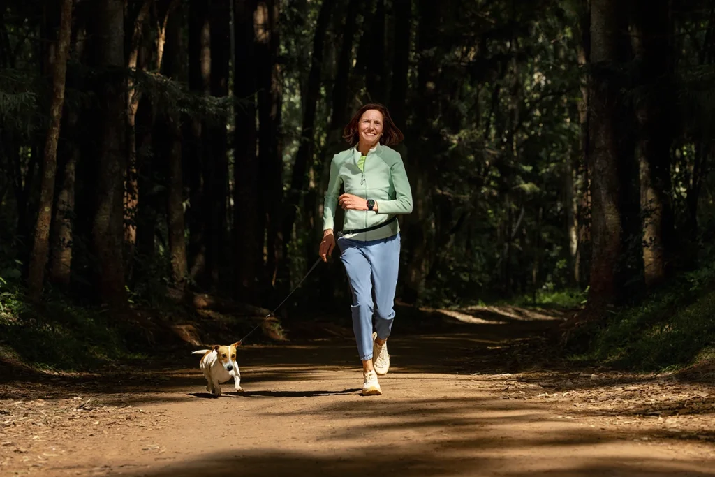 Woman jogging on a dirt forest trail with a small dog on a leash and Watch GT Runner 2 on her hand
