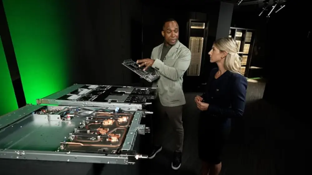 Two professionals examine an open computer chassis, one holding a circuit board as the other watches in a dim lab with a green wall behind them.